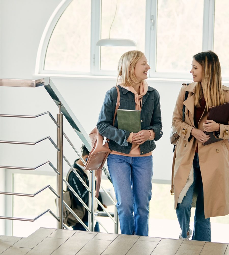 Happy female friends talking while walking in a hallway at the university.